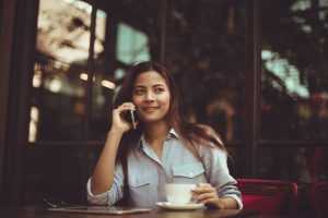 Young woman talking on the phone while sitting at a restaurant drinking coffee, asking about moving services NJ.