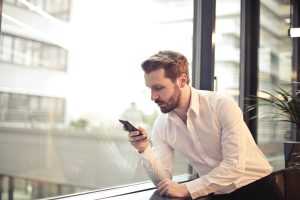 A man standing by the window, following the move on his cell phone.
