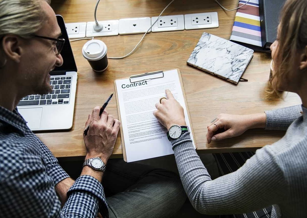 Two people sitting, reading a contract; on the table there is a document titled "Contract", a laptop, coffee, and many sockets