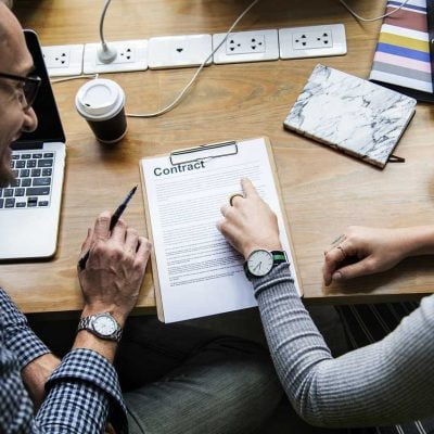 Two people sitting, reading a contract; on the table there is a document titled "Contract", a laptop, coffee, and many sockets