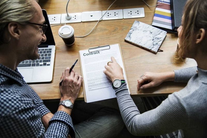 Two people sitting, reading a contract; on the table there is a document titled "Contract", a laptop, coffee, and many sockets