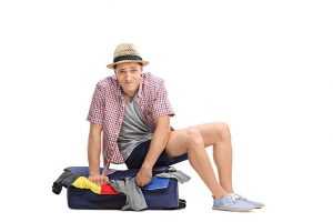A young man sitting on a suitcase full of clothes, trying to close it
