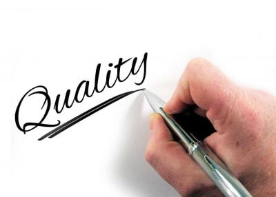 A hand writing the word quality on a white surface
