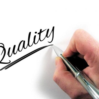 A hand writing the word quality on a white surface